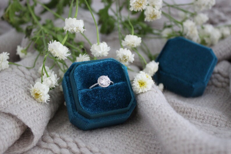 May include: A teal velvet ring box with a diamond engagement ring inside. The box is sitting on a white fuzzy surface with white flowers.