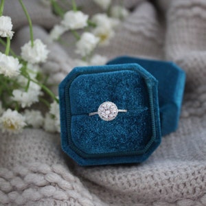 May include: A silver engagement ring with a round diamond center stone and a halo of smaller diamonds, displayed in a teal velvet ring box.