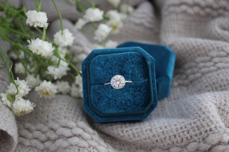May include: A silver engagement ring with a round diamond center stone and a halo of smaller diamonds, displayed in a teal velvet ring box.