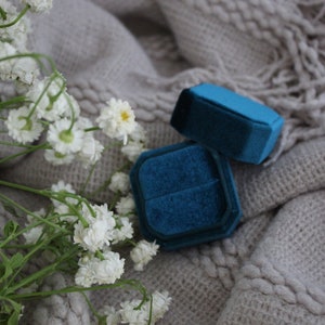 May include: Two blue velvet ring boxes on a white knitted surface with white flowers.