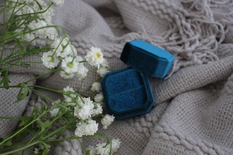 May include: Two blue velvet ring boxes on a white knitted surface with white flowers.