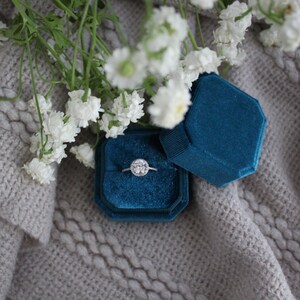 May include: A blue velvet ring box with a silver ring with a round diamond in the center. The box is sitting on a gray knitted surface with white flowers.