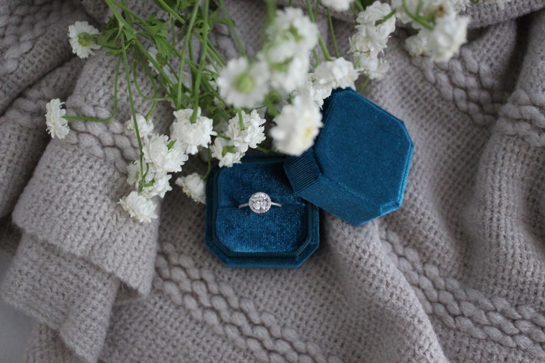 May include: A blue velvet ring box with a silver ring with a round diamond in the center. The box is sitting on a gray knitted surface with white flowers.