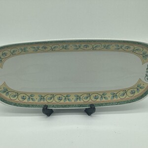 May include: An elongated oval ceramic serving platter featuring a white center and a decorative border. The border displays a pattern of green leaves and small blue flowers on a light tan background, framed by a dark green textured outer edge.