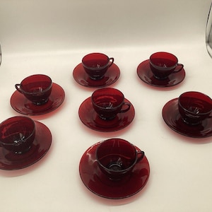 May include: A set of six matching ruby red glass teacups and saucers. Each cup features a rounded bowl shape with a small handle, paired with a circular saucer. The glassware has a deep, translucent red color and a smooth, polished finish.