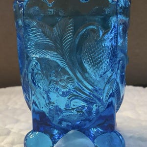 May include: A vibrant blue glass toothpick holder featuring a scalloped top edge and three rounded feet. The exterior surface is decorated with a raised, embossed pattern of detailed leaves and flowers, creating a textured finish on the translucent glass.