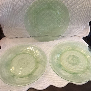 May include: Three light green glass plates featuring a textured diamond pattern across the surface and a decorative embossed floral motif in the center. The plates have a translucent appearance and a circular shape with a smooth, slightly raised rim.
