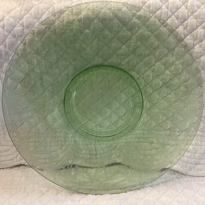 May include: A round, light green glass plate featuring a central circular depression and a wide, flat rim. The surface is decorated with an embossed, repeating diamond-shaped lattice pattern and intricate, stylized floral motifs along the outer edge.