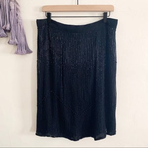 May include: A black knee-length skirt with vertical beaded embellishments. The skirt hangs from a black waistband and features a subtle chevron pattern created by the beading. The skirt is displayed on a wooden hanger.