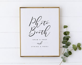 May include: A gold-framed sign with the text "Photo Booth" in a black cursive font. The sign also includes the text "Grab a prop and strike a pose!" in a smaller, black font.