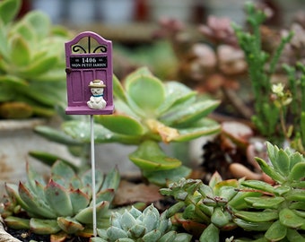 May include: A miniature purple door with the text "1406 MON PETIT JARDIN" and a small figurine of a cat wearing a hat. The door is on a metal stake, placed among green succulents.
