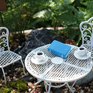 May include: A miniature white metal patio set with two chairs and a table. The table has a blue book and two white cups with coffee on it.