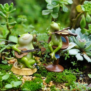 May include: Two green frog figurines, one playing a guitar and the other playing a violin, sitting on a mossy surface with green plants.