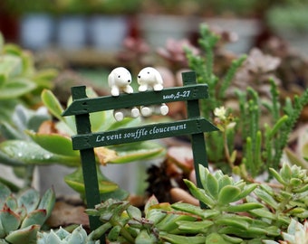 May include: Two white ceramic dog figurines sitting on a green wooden sign with the text "Le vent souffle clousement" and "rue, 5-27". The sign is surrounded by green succulents.