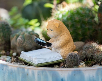 May include: A small, tan figurine of a dog wearing a magnifying glass and looking at an open book. The figurine is sitting on a bed of small cacti and rocks.