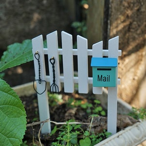 Peut inclure: Une petite clôture en bois blanc avec une petite boîte aux lettres bleue sur laquelle est inscrit "Mail". Deux petits outils de jardinage sont accrochés à la clôture, une pelle et un râteau.