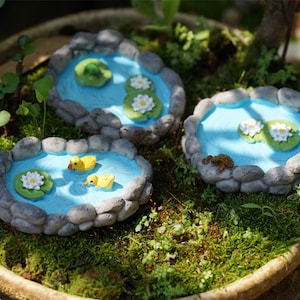 Puede incluir: Tres estanques de piedra en miniatura con agua azul, nenúfares y flores blancas. Un estanque tiene dos figuras de patos amarillos nadando en el agua.