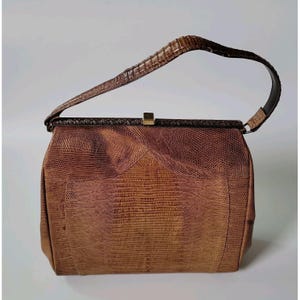 Può includere: Una borsa a mano strutturata di colore marrone con una trama testurizzata effetto pelle di lucertola. La borsa presenta un manico arcuato, una struttura rettangolare e una piccola chiusura metallica dorata al centro. L'esterno mostra una sfumatura di toni marroni.