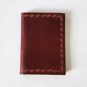 May include: A rectangular dark brown leather cover featuring a decorative border of embossed scrollwork patterns along the edges. The leather has a smooth, matte finish with visible stitching along the perimeter of the front panel.