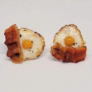 Può includere: Un paio di orecchini a forma di uova fritte con pancetta. Le uova sono bianche con un tuorlo giallo e pepe nero. La pancetta è marrone e croccante.