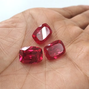May include: Three red cushion-cut gemstones, likely rubies, are displayed on a hand. The gemstones are faceted and have a high degree of brilliance.