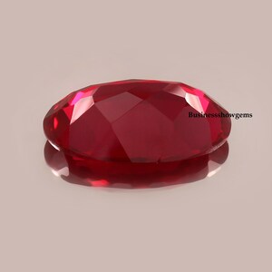 May include: A deep red oval-shaped gemstone with a faceted cut. The gemstone is reflecting light and has a shiny finish.