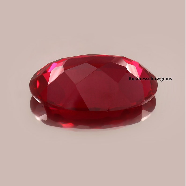 May include: A deep red oval-shaped gemstone with a faceted cut. The gemstone is reflecting light and has a shiny finish.