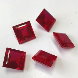 May include: Five red square-shaped gemstones with a faceted cut. The gemstones are arranged on a white surface.