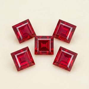 May include: Five square-cut, deep red gemstones arranged in a cluster. Each stone features a faceted surface with a pointed center, reflecting light to create a geometric pattern of dark and bright crimson tones across the transparent surfaces.