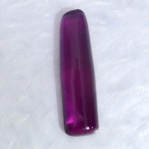 May include: A single, elongated, rectangular-shaped gemstone with a smooth, polished surface and a deep, translucent purple color. The object features rounded corners and a uniform, solid violet hue throughout its entire length and width.