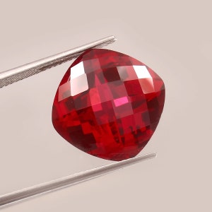 May include: A deep red gemstone with a cushion cut and many facets. The gemstone is held by a pair of tweezers.