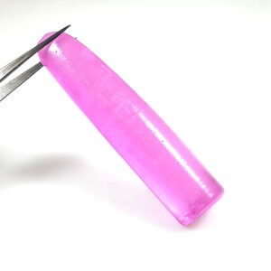 May include: A vibrant, elongated, pink gemstone is held by silver tweezers against a white background. The gemstone has a smooth, polished surface and a rectangular shape with rounded edges. The color is a bright, saturated pink.
