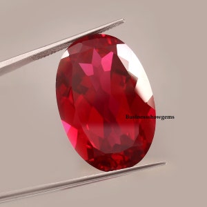 May include: A deep red oval-shaped gemstone with a faceted cut. The gemstone is held by tweezers against a white background.
