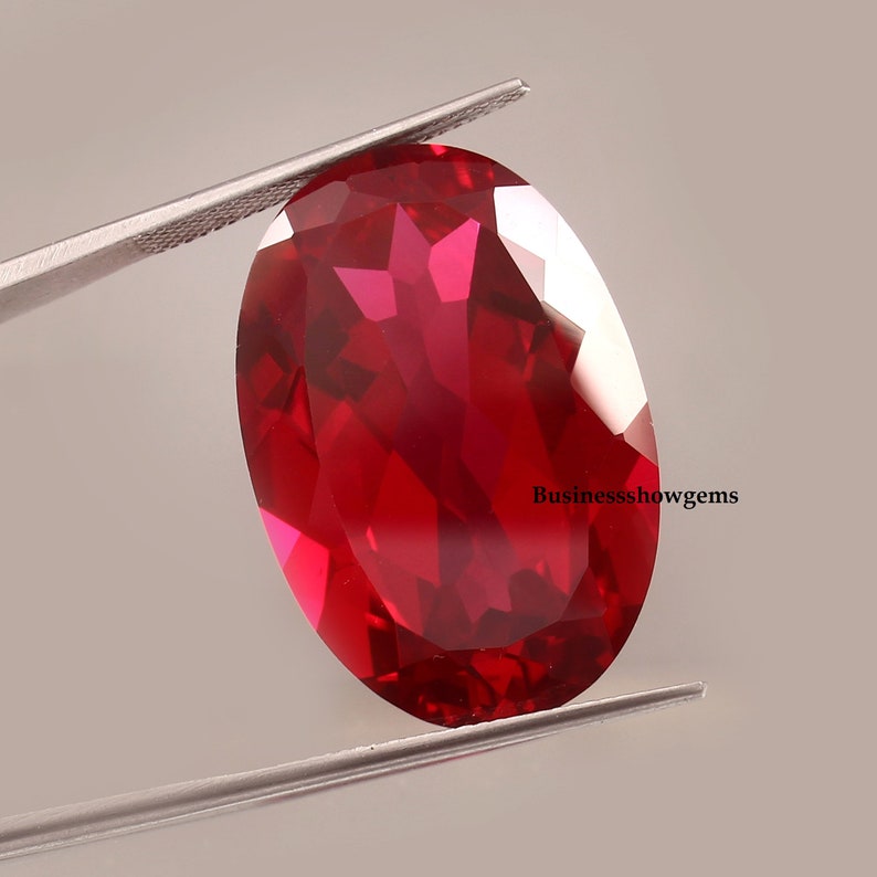 May include: A deep red oval-shaped gemstone with a faceted cut. The gemstone is held by tweezers against a white background.