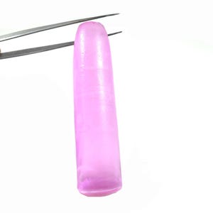 May include: A long, slender, translucent pink gemstone held by tweezers. The gemstone is smooth and polished, with a slight shimmer.