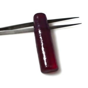 Può includere: Una pietra preziosa sfaccettata di colore rosso intenso tenuta da una pinzetta. La pietra preziosa è lunga e stretta, con una superficie liscia e lucida.