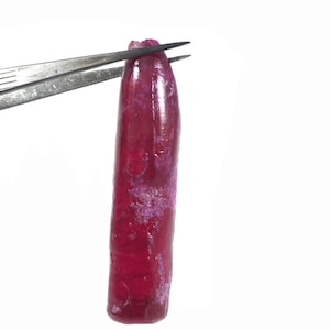 May include: A raw, elongated ruby gemstone, held by silver tweezers against a white background. The ruby is a deep, saturated red with some lighter, purplish areas. The gemstone has a rough, natural texture.