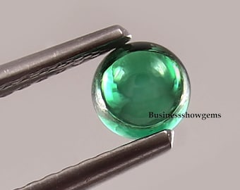 Puede incluir: Una piedra preciosa redonda y verde con una superficie lisa y pulida. La piedra preciosa se sostiene con unas pinzas.