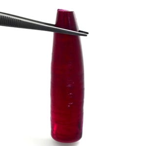 May include: A deep red, translucent, elongated gemstone with a smooth, cylindrical shape and a slightly tapered top. The stone exhibits a rich, saturated crimson hue throughout its body, with subtle internal variations in light transmission.