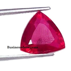 May include: A triangular, faceted ruby gemstone with a deep red color. The ruby is held in tweezers against a white background.