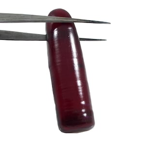 Può includere: Una gemma allungata di colore rosso intenso tenuta da una pinzetta. La gemma è liscia e lucida, con un leggero bagliore.