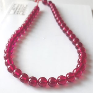 Puede incluir: Una hebra de cuentas redondas de color rojo intenso, enhebradas para formar un collar. Las cuentas son lisas y brillantes, con un ligero brillo.