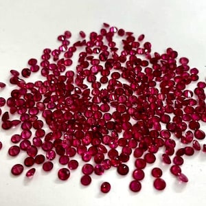 May include: A large collection of small, round, faceted red rubies scattered across a plain surface. Each gemstone displays a brilliant crimson hue with multiple facets that catch the light, creating a sparkling effect across the entire group.