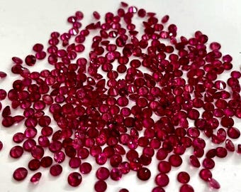 Puede incluir: Una gran colección de pequeños rubíes rojos redondos y facetados esparcidos sobre una superficie lisa. Cada piedra preciosa muestra un tono carmesí brillante con múltiples facetas que captan la luz, creando un efecto centelleante.
