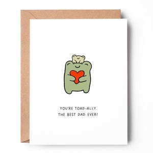 May include: A white greeting card with a brown envelope featuring a cartoon illustration of a green frog holding a red heart with a small beige frog on its head. The text below the illustration reads, "YOU'RE TOAD-ALLY THE BEST DAD EVER!"