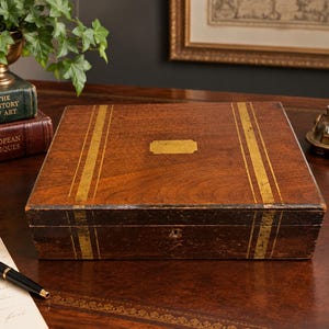 May include: A rectangular wooden box with a dark brown finish, featuring two vertical gold-colored stripes on the lid and a central rectangular brass-colored plaque. The box has a keyhole on the front side and a smooth, polished wood grain surface.