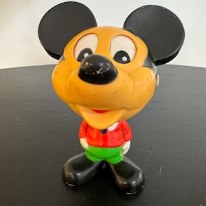 May include: A vintage Mickey Mouse figurine with a yellow head, black ears, and a red shirt. The figurine has green shorts and black shoes. The figure is standing on a black surface.