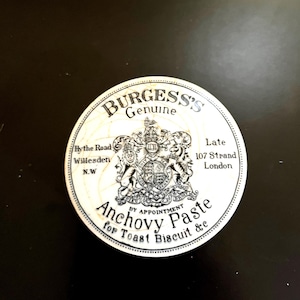May include: A white round jar lid with black text and a crest. The text reads "Burgess's Genuine", "By the Road Willesden N.W", "Late 107 Strand London", "By Appointment", "Anchovy Paste for Toast Biscuit &c".