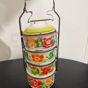 May include: Vintage metal tiffin carrier with three stacked compartments. Each section is white with a floral design in red, orange, and yellow. The carrier has a metal frame and a wooden handle. A charming, decorative piece.