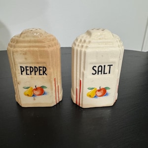 May include: A pair of vintage ceramic salt and pepper shakers. The pepper shaker is tan and the salt shaker is white. Each shaker has a fruit design and the words "PEPPER" and "SALT" in black lettering.